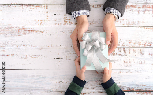 Closeup & Top view, a little child's hands hand over a beautiful gift, glittering blue wrap and big bow, to dad in business suit on wooden table. Thankful, grateful and happy father's day concept.
