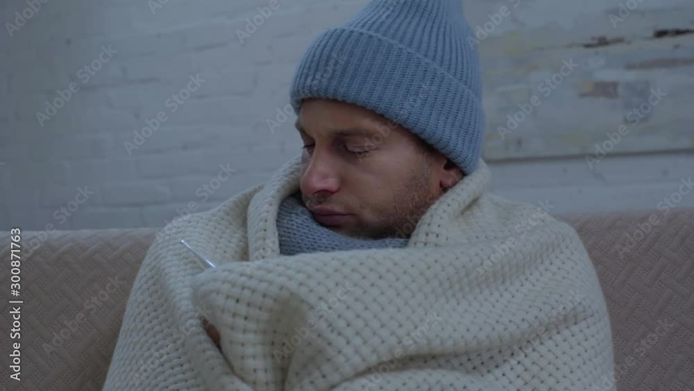 sick man in warm hat wrapping in blanket and measuring temperature