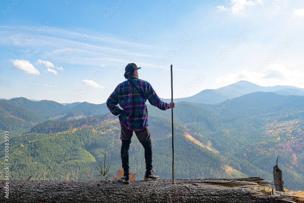 Naklejka premium Young male traveler with stick in hands standing on a big log, mountains and sky on background