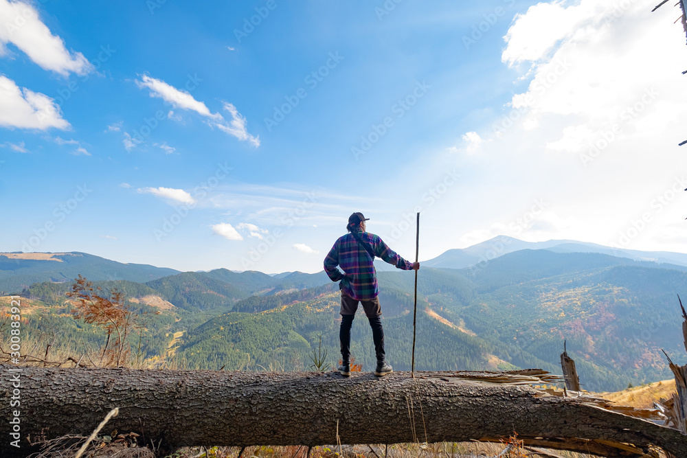 Naklejka premium Young male traveler with stick in hands standing on a big log, mountains and sky on background