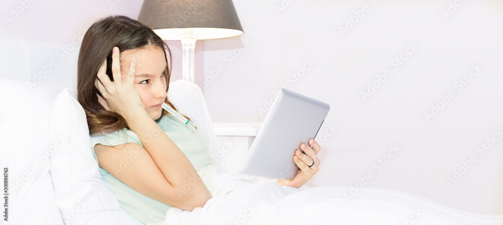 Beautiful teenage girl looking and using tablet while lying in comfort bed in the morning in bedroom entertaining with mobile apps Beautiful teenage girl looking and using tablet while lying in comfort bed in the morning in bedroom entertaining with mobile apps