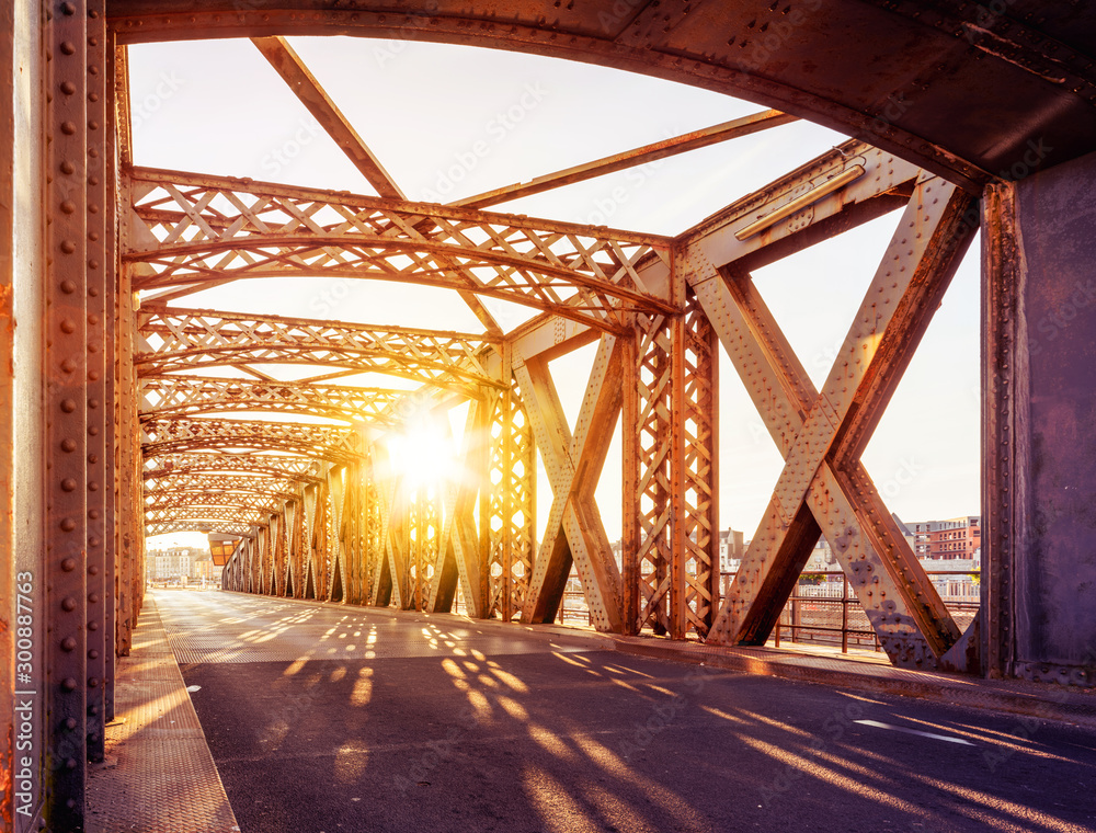 Asphalt road under the steel construction of a bridge in the city on a sunny day. Evening urban scene with the sunbeam in the tunnel. City life, transport and traffic concept. Asphalt road under the steel construction of a bridge in the city on a sunny day. Evening urban scene with the sunbeam in the tunnel. City life, transport and traffic concept.