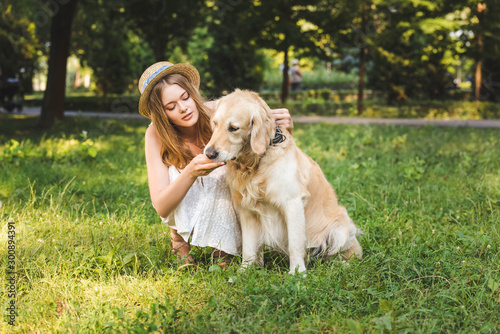 full length view of beautiful girl in white dress and straw hat petting golden retriever while sitting on meadow and looking at dog