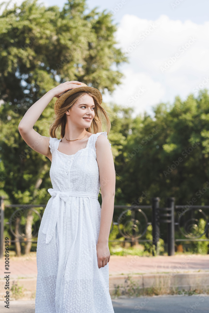 beautiful young girl in white dress touching straw hat while smiling and looking away beautiful young girl in white dress touching straw hat while smiling and looking away