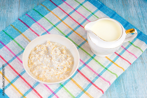 Simple breakfast - cup with oatmeal, milk jug with milk on a napkin on a wooden table in blue tones close up