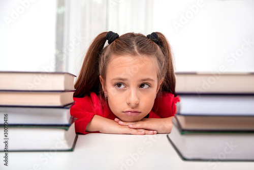 Shot of young girl   sitting at  the table with two large stacks of books and   looking dreamily into the distance.