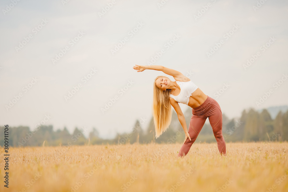 Beautiful blonde girl in white T-shirt is stretching in park field, yoga concept - obrazy, fototapety, plakaty 