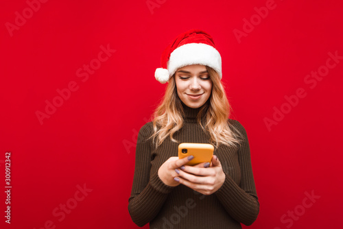 Positive lady in christmas hat uses smartphone on red background  looks at screen and smiles. Cute girl in santa claus hat uses internet on phone  isolated.