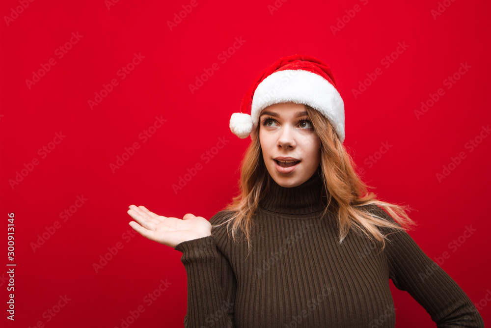 Astonished attractive girl in warm sweater on christmas hat santa claus looks and points with her hand to the empty space, isolated. Emotional girl holding copyspace in her hand on red background. Astonished attractive girl in warm sweater on christmas hat santa claus looks and points with her hand to the empty space, isolated. Emotional girl holding copyspace in her hand on red background.