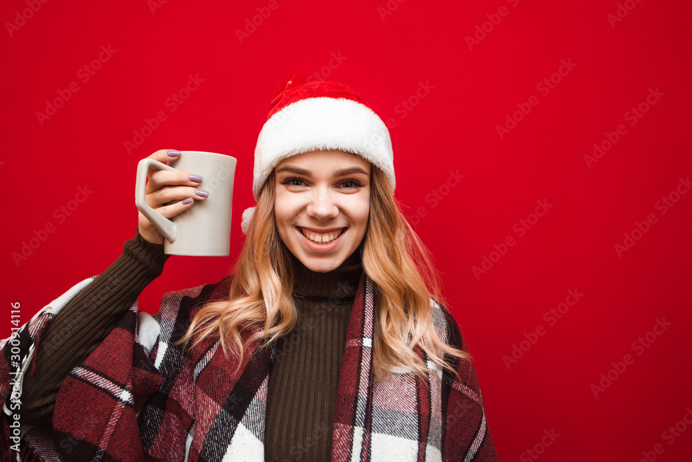 Christmas portrait of happy girl in plaid and santa hat on red background, holds big cup in hand with warm drink, looks into camera and smiles. Joyful girl drinking tea at Christmas, isolated. Christmas portrait of happy girl in plaid and santa hat on red background, holds big cup in hand with warm drink, looks into camera and smiles. Joyful girl drinking tea at Christmas, isolated.