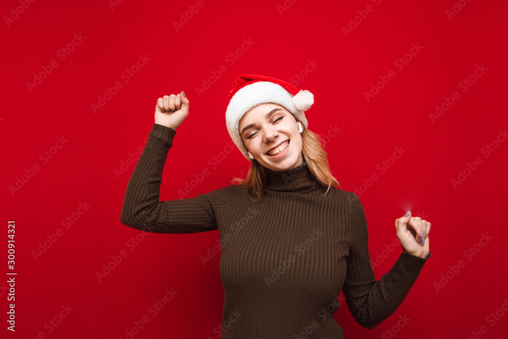 Happy girl in santa hat and warm sweater listens to music in closed-loop headphones, smiles and dances on red background. Christmas party for music in headphones. Xmas Happy girl in santa hat and warm sweater listens to music in closed-loop headphones, smiles and dances on red background. Christmas party for music in headphones. Xmas