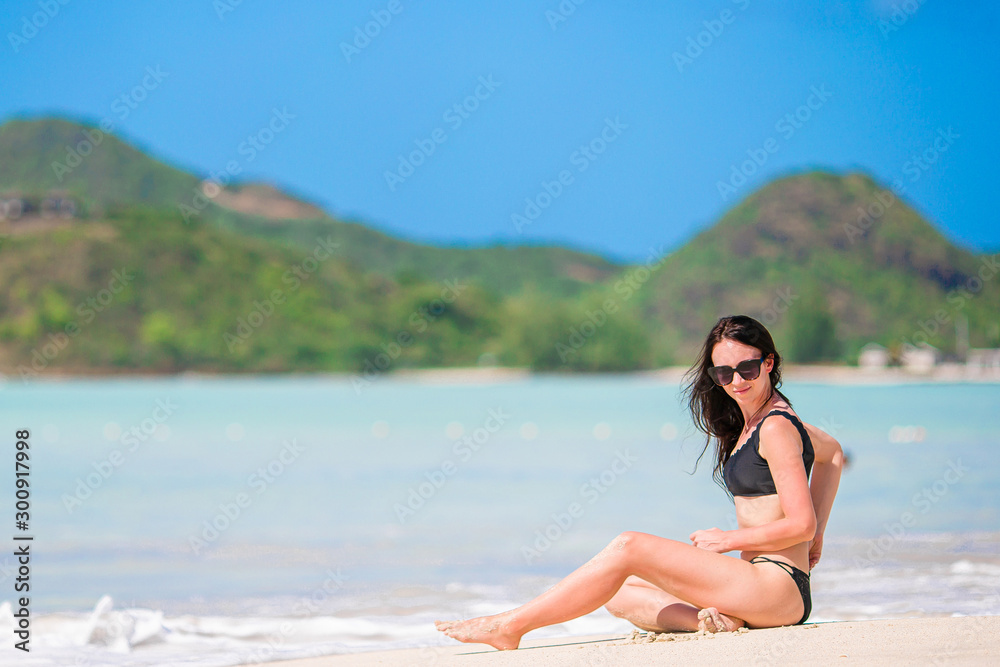Young happy woman in swimsuit on white beach. Beautiful model in bikini sitting down.