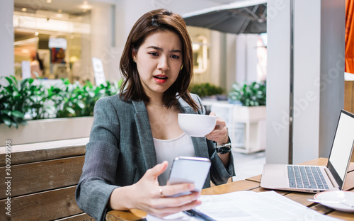 Confused young Asian businesswoman with coffee cup using smartphone