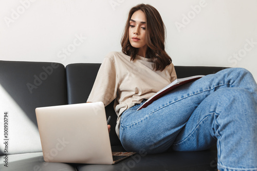 Woman sit indoors at home using laptop holding copybook.
