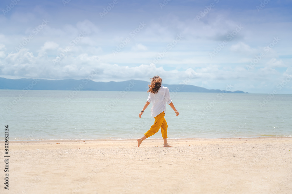 Young beautiful woman in a white shirt and yellow pants runs joyfully on the beach along the sandy shore of a tropical sea