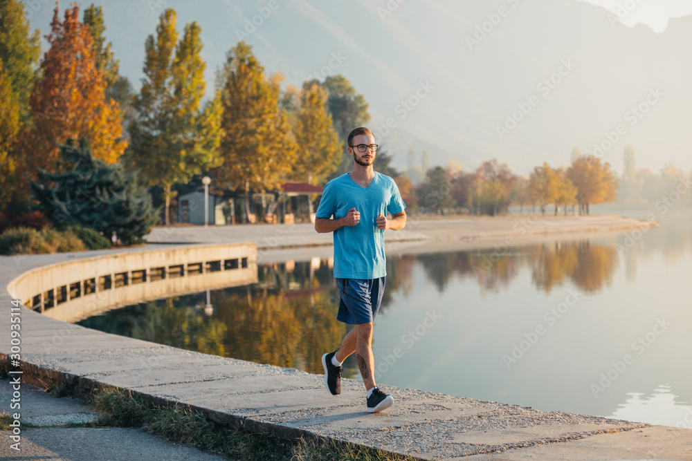Young man with glasses in sportswear jogging by the lake in a city park. Runner training in the park by the water at sunset.