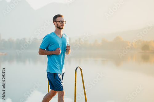 Young man with glasses in sportswear jogging by the lake in a city park. Runner training in the park by the water at sunset. © Suteren Studio