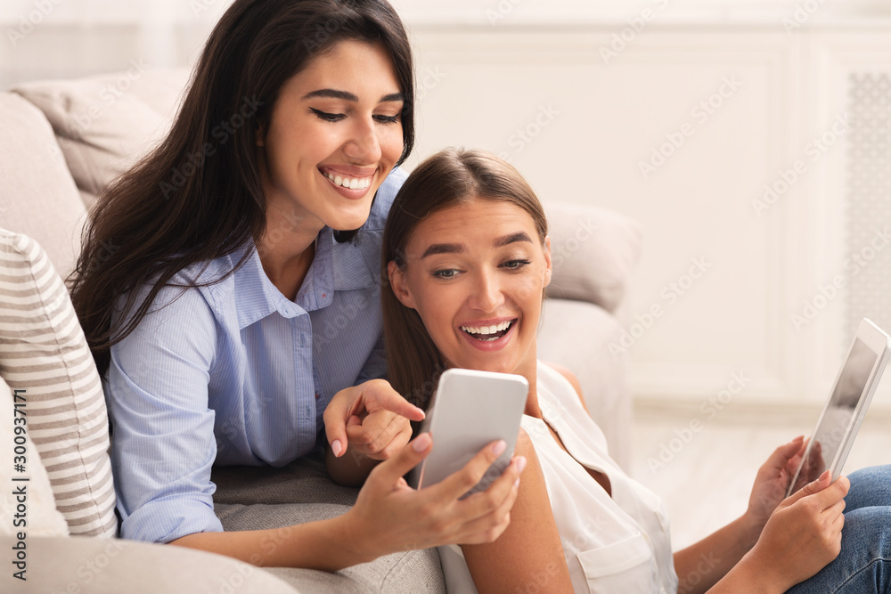 Two Girls Using Phone And Tablet Sitting On Sofa Indoor