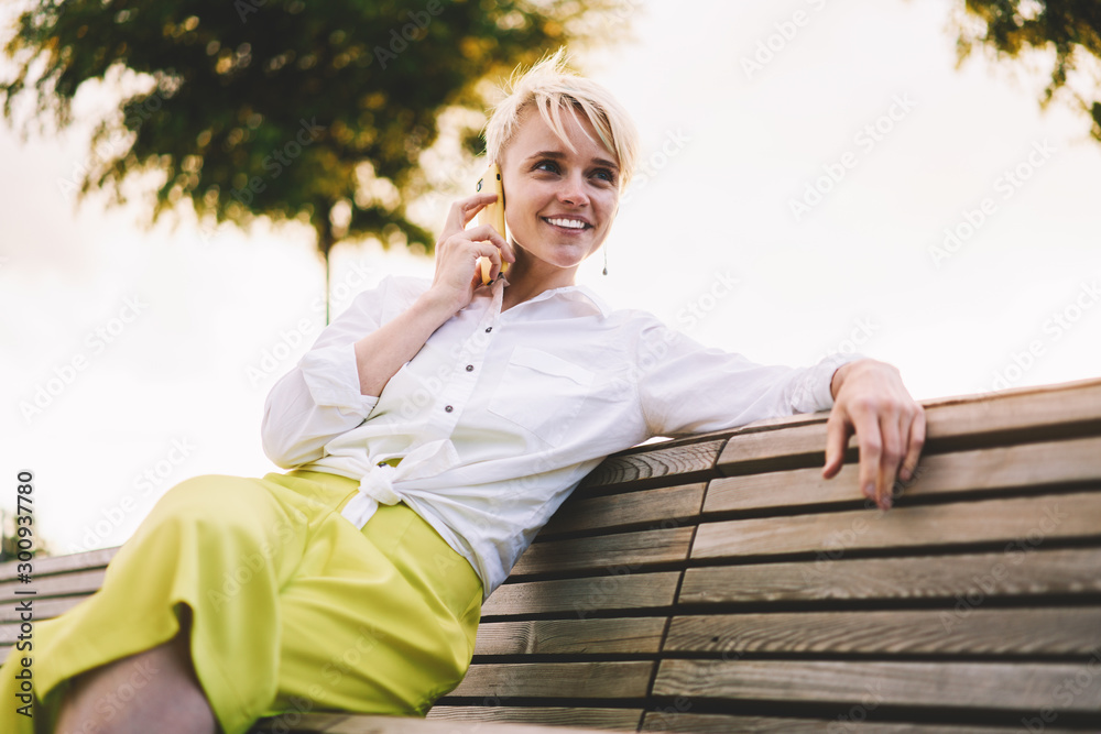 Caucasian cheerful woman in trendy outfit smiling and enjoying sunny weather in park while communicating with best friend via cellular gadget using roaming connection for international calls Caucasian cheerful woman in trendy outfit smiling and enjoying sunny weather in park while communicating with best friend via cellular gadget using roaming connection for international calls