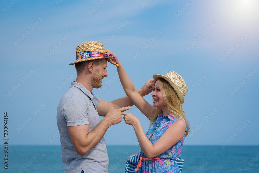 Cute European loving couple in sun hats having fun on the pier near clear blue sea, they smiling, pointing to each other and looking to each other. They are on their honeymoon.