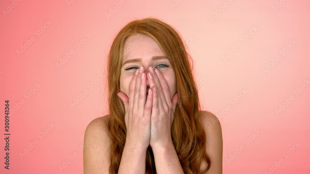 In front of a bright pink backdrop, a young woman breaks into laughter, holding her hands over her mouth In front of a bright pink backdrop, a young woman breaks into laughter, holding her hands over her mouth