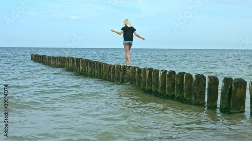 A young blonde woman walks carefully along some wooden posts in shallow water on a beach