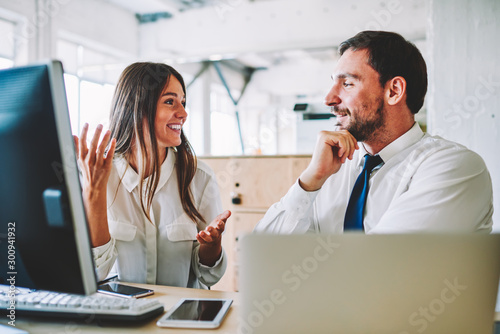 Pensive bearded male employee carefully listening attractive smiling female co-worker during collaboration in modern office.Positive man and woman graphic designers communicating in good atmosphere