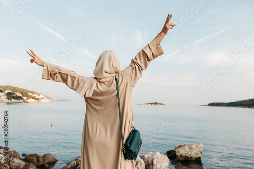 portrait of young European Muslim women with hijab standing on the stone beach with her hand s in the air. Sea is in the background. She is happy and relaxed. photo