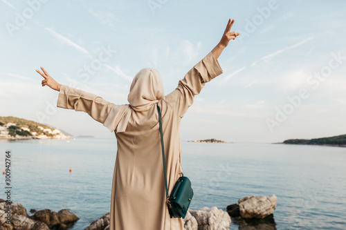 portrait of young European Muslim women with hijab standing on the stone beach with her hand s in the air. Sea is in the background. She is happy and relaxed.