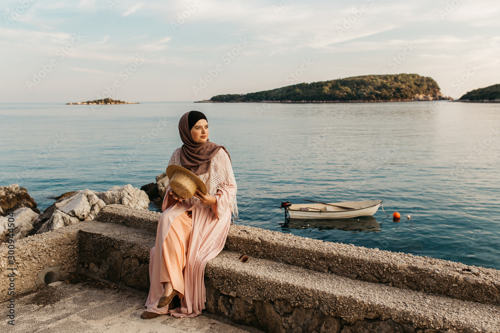 portrait of young European Muslim women with hijab sitting on the beach with hat in her hand. She is happy and relaxed. Sea is in the background. Looking in the distance. portrait of young European Muslim women with hijab sitting on the beach with hat in her hand. She is happy and relaxed. Sea is in the background. Looking in the distance.