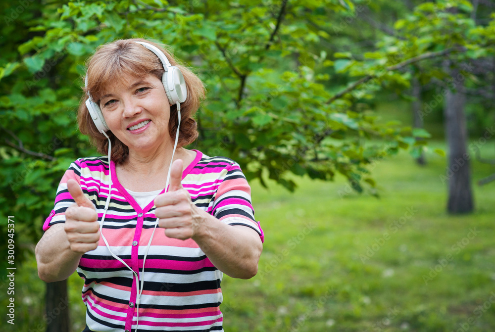 Beautiful joyful middle aged European woman in white headphones shows thumbs up in park in summer Beautiful joyful middle aged European woman in white headphones shows thumbs up in park in summer