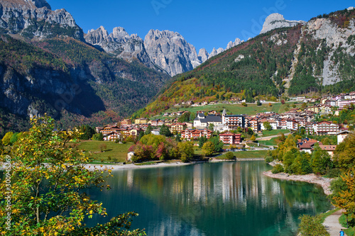 Fototapeta Naklejka Na Ścianę i Meble -  Aerial view of Lake Molveno, north of Italy in the background the city of Molveno, Alps, blue sky. Autumn season. Multi-colored palette of colors