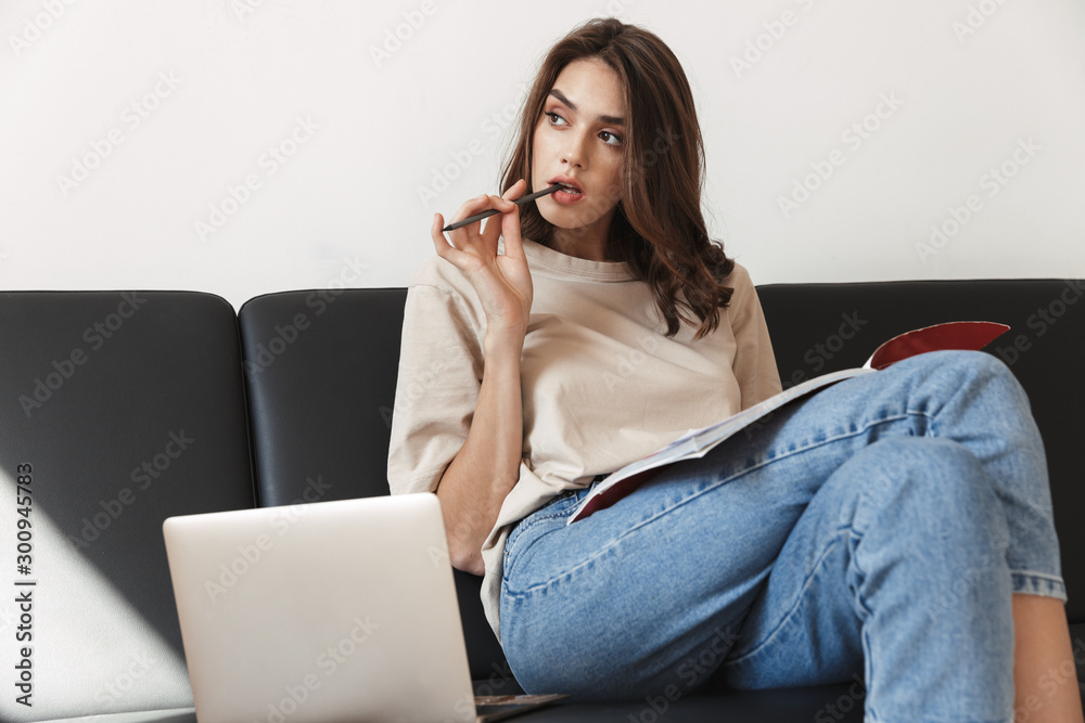 Image of woman studying with exercise book and laptop on sofa in apartment
