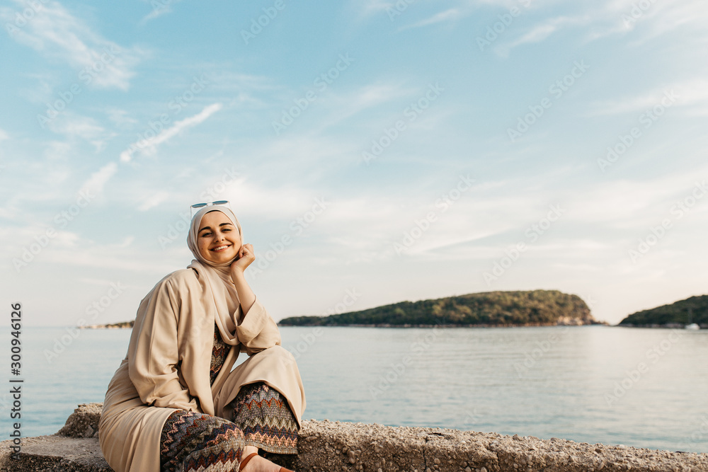 portrait of young European Muslim women with hijab sitting on the stone beach with sea in the background. She is happy and relaxed.