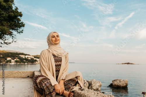 portrait of young European Muslim women with hijab sitting on the stone beach with sea in the background. She is happy and relaxed.