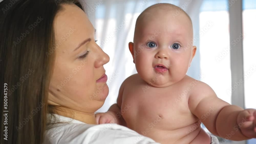 Mother holds a chubby smiling baby in her arms at home. The kid is looking at the camera. Close up shot. Mother holds a chubby smiling baby in her arms at home. The kid is looking at the camera. Close up shot.