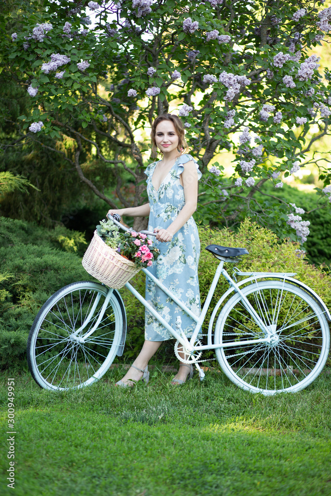 portrait of a beautiful girl in the forest, holding a bike with a basket of flowers, behind the rays of the sun, a blue flowered dress, summer walk