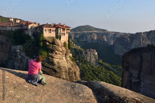 Sitting on the rock woman takes photo of Varlaam Monastery at sunset, Rosanou nunnery seen at distance. Meteora - UNESCO world heritage site, Greece 