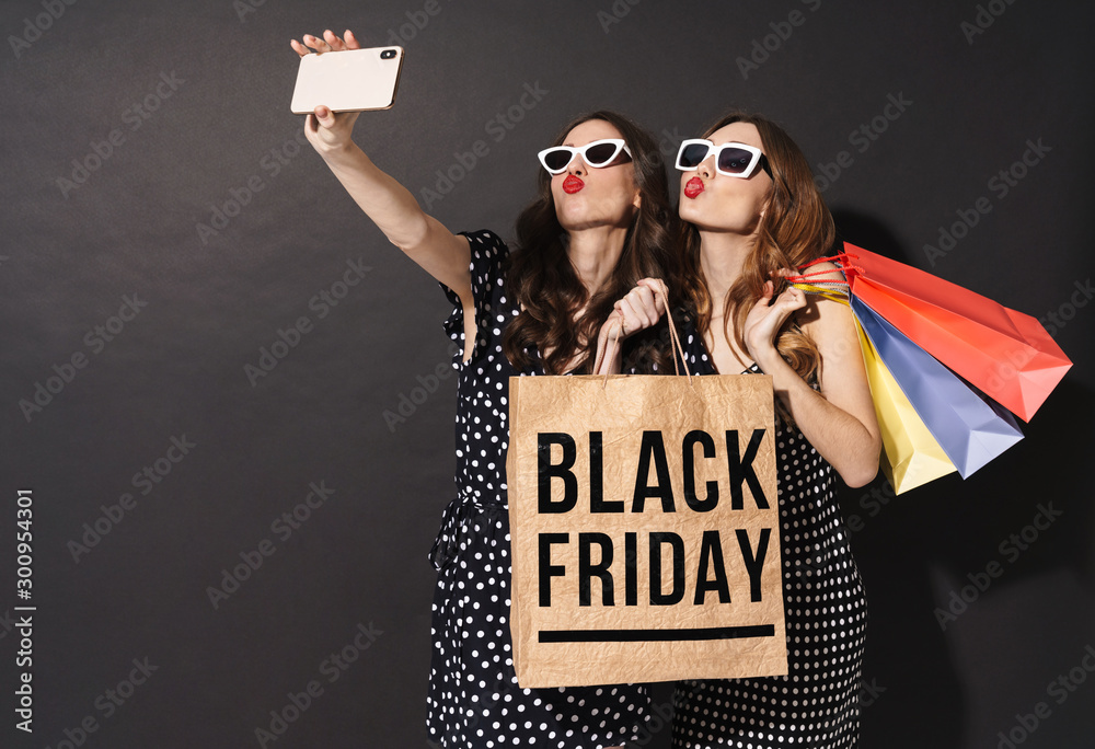 Image of girls taking selfie on cellphones and holding black friday bag - obrazy, fototapety, plakaty 
