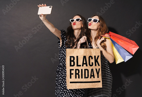 Image of girls taking selfie on cellphones and holding black friday bag