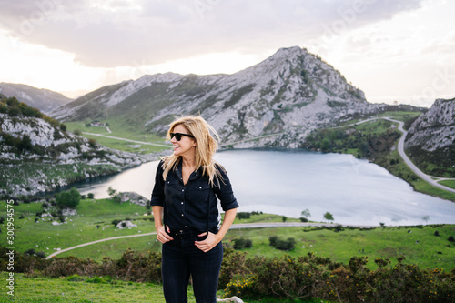 A beautiful caucasian blond woman in front of the camera wearing casual clothes in a mountainous landscape with lake