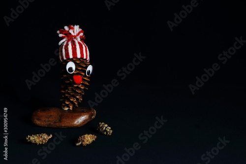 Cheerful homemade New Year man made of cones in bright decorative hat on wooden stand and cones on black background. Symbolic concept fantasy, imagination, New Year, holiday. Minimal style.