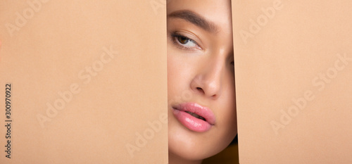 Lady with nude makeup peering into hole in peach paper © Prostock-studio