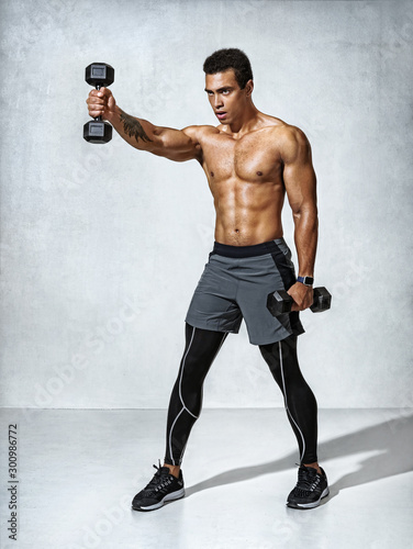 Young man bodybuilder doing exercise with heavy weight dumbbells. Photo of muscular man on grey background. Strength and motivation.