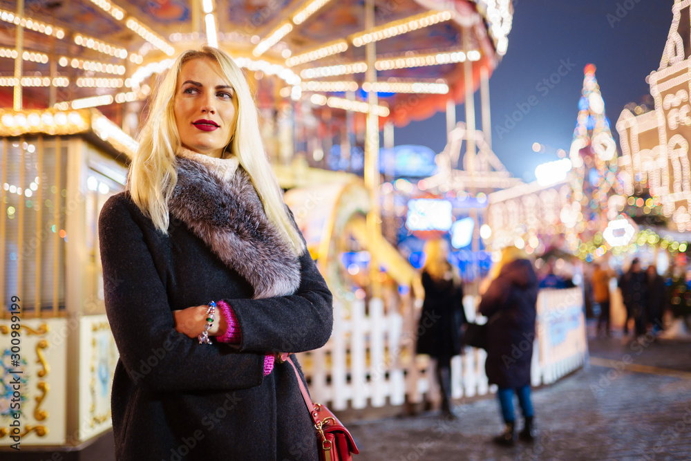 Picture of young woman in coat with fur collar in park on background of carousel