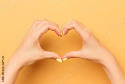 Female hands show a heart symbol isolated. Bright manicure. Warm yellow background