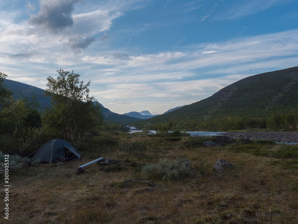 Small green tent in Beautiful wild Lapland nature landscape with blue river, Kaitumjaure lake, birch trees and mountains. Northern Sweden summer at Kungsleden hiking trail. Blue sky dramatic clouds
