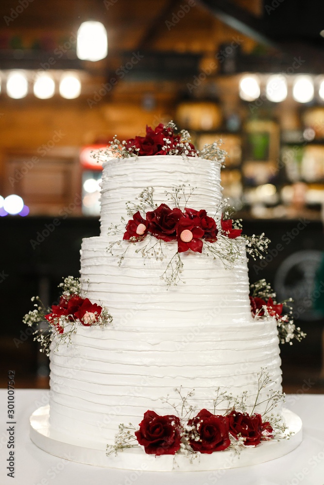  white three-tiered wedding cake  standing on the table. festive cake decorated with mastic and flowers on the holiday table . beautiful white high birthday cake with red flowers