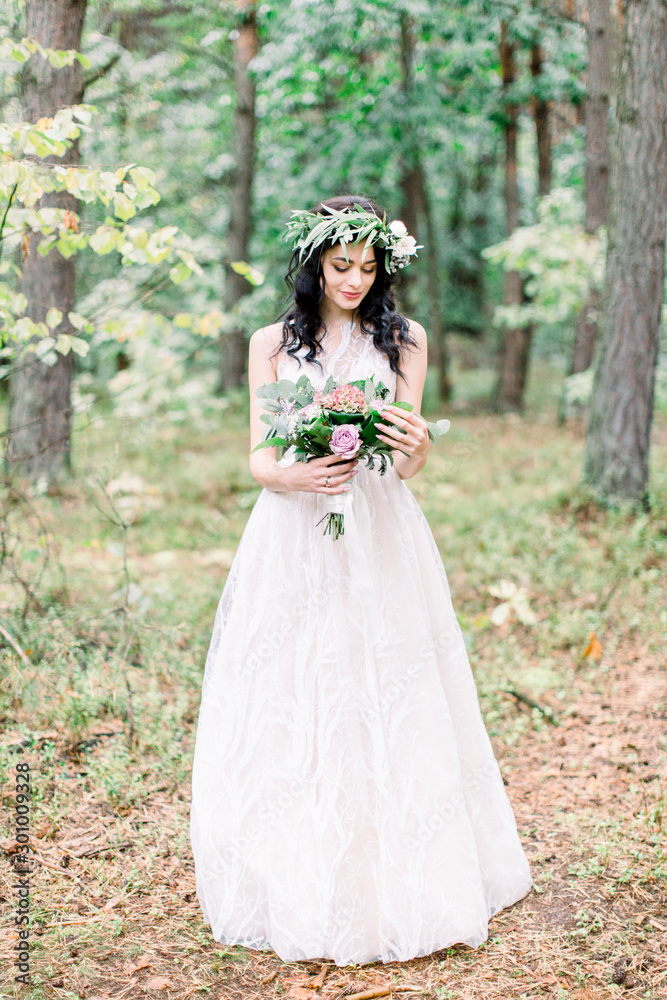 Beautiful bride with eucalypthus wreath on the head standing in the forest and looking at wedding bouquet of different flowers. Rustic style