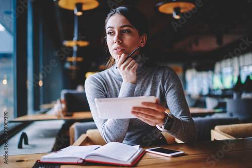 Caucasian brunette woman thoughtfully looking aside while resting in cafe with new gadget, attractive dreamy hipster girl planning tomorrow list using portable pc app during break from work indoors photo